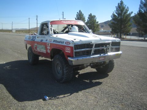 1990 Ford Bronco - red-and-white off-road race truck numbered 302 with tube bumper and BFGoodrich graphics sits on a gravel lot under blue sky.