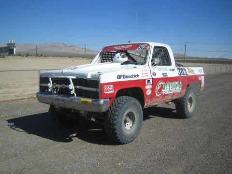 1990 Ford Bronco - red-and-white off-road race truck numbered 302 with California Trophy and BFGoodrich livery parked on a desert gravel lot.