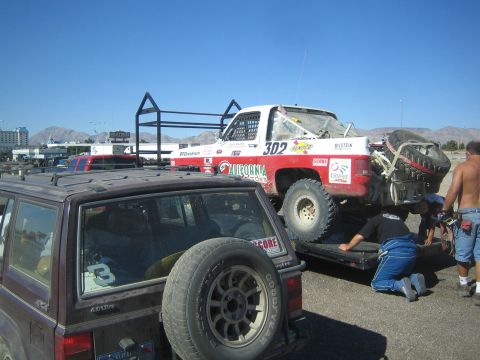 1990 Ford Bronco - red-and-white number 302 off-road race truck sits on a flatbed trailer in a pit area while a crew works on a nearby vehicle.