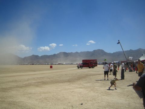 1990 Ford Bronco - Crowds gather on a dusty open desert lakebed with mountains behind as a red box truck and a camera boom stand in the distance.