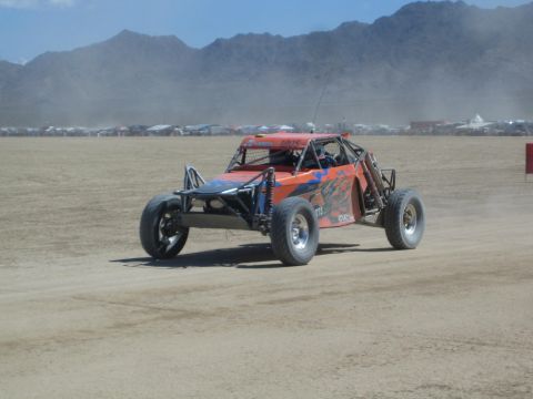 1990 Ford Bronco - orange tube-frame off-road racing buggy speeds across a dusty desert course with mountains and spectator camps behind.