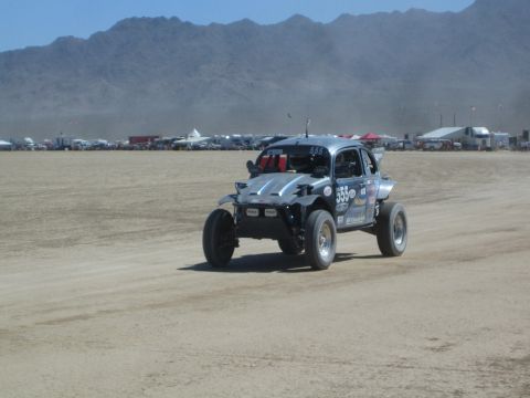 1990 Ford Bronco - silver Baja-style off-road buggy with sponsor decals races across a dusty desert flat lined with distant camps and mountains.
