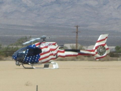 1990 Ford Bronco - helicopter painted with a stars-and-stripes American flag scheme, registration N702, rests on a desert flat with mountains behind.