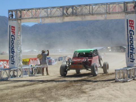 1990 Ford Bronco - red off-road buggy with a green roll-cage top passes under a BFGoodrich banner arch as an official waves on a dusty desert course.