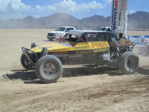 1990 Ford Bronco - yellow tube-frame off-road buggy numbered 1212 with Curb Craters sponsor decals races on a dusty desert course.