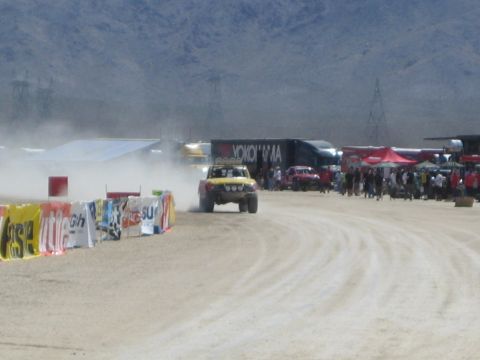 1990 Ford Bronco - off-road race truck kicks up a dust cloud rounding a desert course past colorful sponsor banners and parked transporters.
