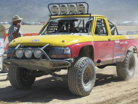 1990 Ford Bronco - yellow and pink off-road race truck topped with four Hella driving lights sits on a dusty desert course as an official watches.
