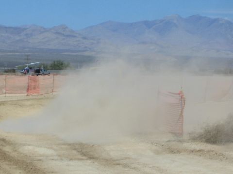 1990 Ford Bronco - thick cloud of dust obscures a desert race course lined with orange snow fencing, with mountains visible on the horizon.