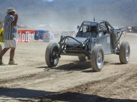 1990 Ford Bronco - dark gray tube-frame off-road buggy numbered 1616 races on a dusty desert course as a striped-shirt official looks on.