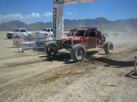 1990 Ford Bronco - orange tube-frame off-road buggy accelerates past a BFGoodrich banner arch on a wide dusty desert flat with mountains behind.
