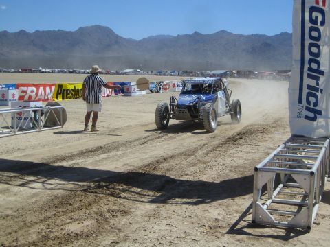 1990 Ford Bronco - blue tube-frame off-road buggy races down a dusty desert course past a BFGoodrich banner as an official watches near the start.