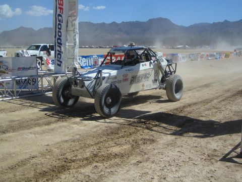 1990 Ford Bronco - white tube-frame off-road buggy with sponsor decals rounds a dusty desert course near BFGoodrich and Suzuki start banners.