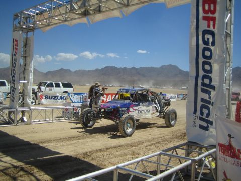 1990 Ford Bronco - open-wheel off-road race buggy passes beneath a BFGoodrich-branded start arch on a dusty desert course.