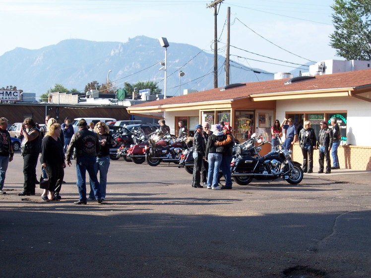 Riders and parked cruiser motorcycles outside a storefront with mountains behind
