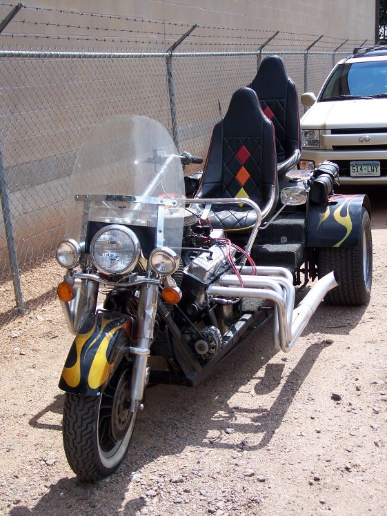 Custom trike with two seats, V8 engine, and flame decals. A light-colored SUV is parked behind it