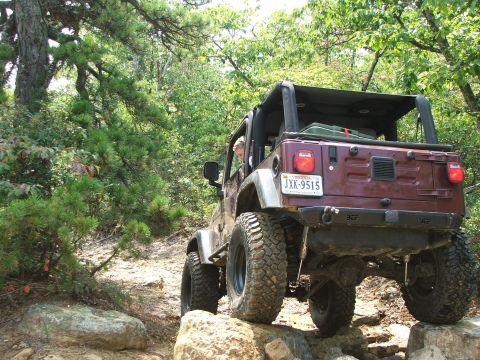 1996 Ford Bronco - A maroon Jeep Wrangler navigates a challenging rocky trail surrounded by trees.