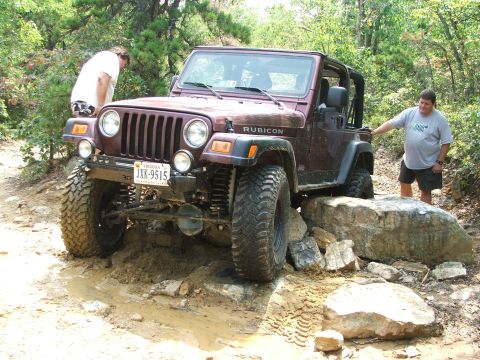 1996 Ford Bronco - A Jeep Wrangler Rubicon tackles a challenging off-road course with large rocks and muddy terrain.