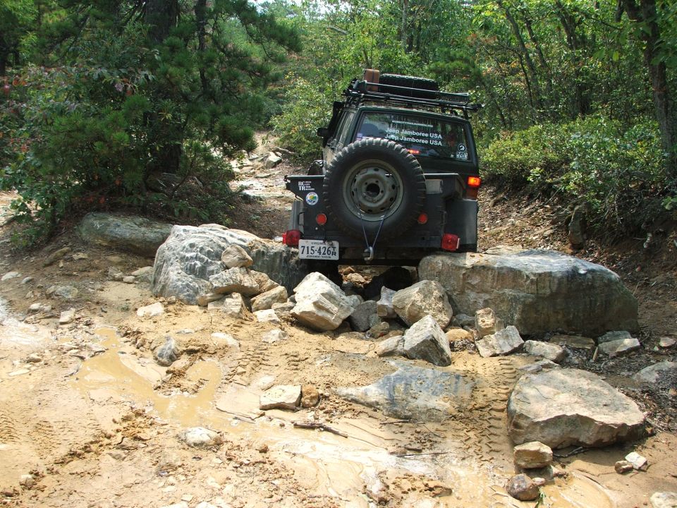 Jeep-like vehicle on rocky trail, spare tire on back, roof rack, license plate 715-426X