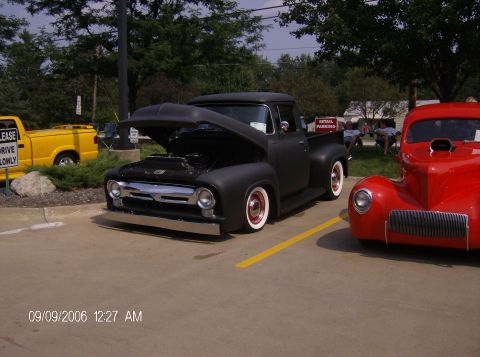 1970 Ford Ranchero - A matte black 1956 Ford F-100 pickup truck is displayed with its hood open, parked next to a red hot rod.