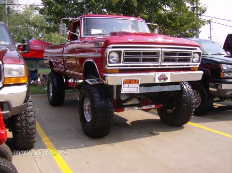 1970 Ford Ranchero - A lifted red pickup truck is parked on display, with a "FOR SALE" sign on the bumper and other vehicles nearby.