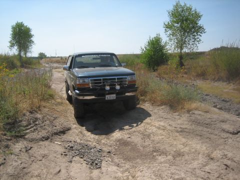 1995 Ford Bronco - This 1995 Ford Bronco is captured in a 3q-front view as it navigates a dusty, unpaved road under a clear blue sky.