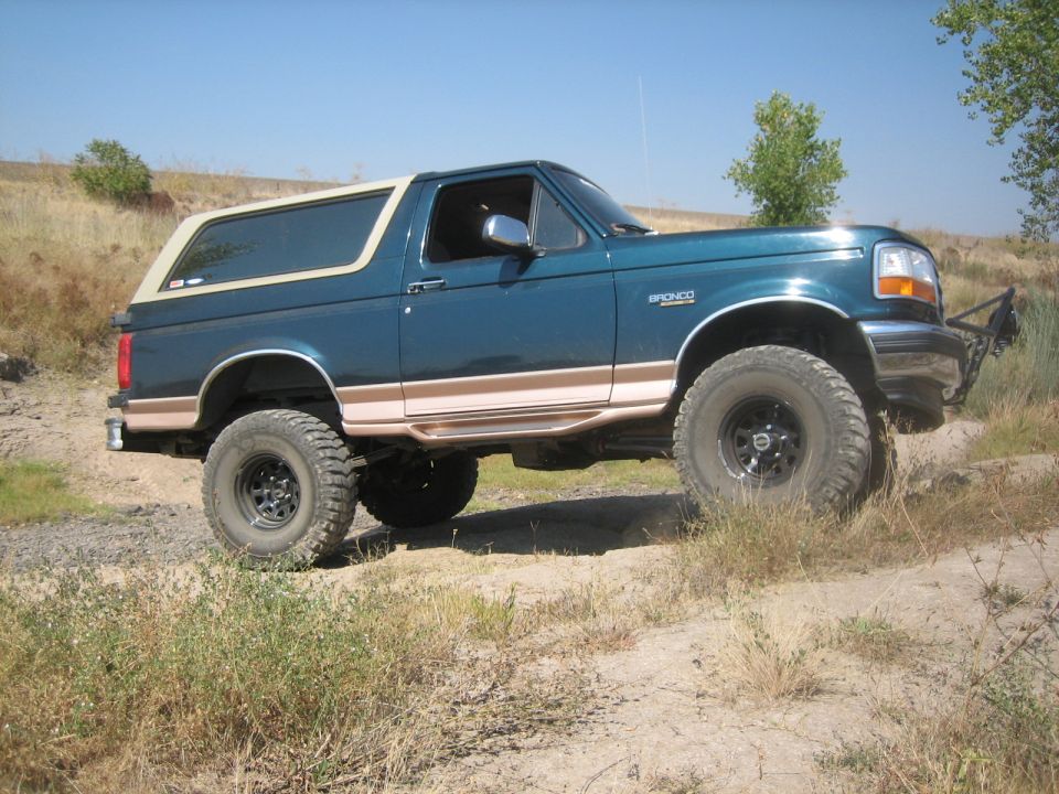 Teal Ford Bronco with tan trim, lifted on off-road tires, driving on a dirt path