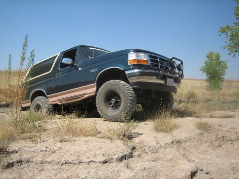 1995 Ford Bronco - A dark green Ford Bronco with tan trim navigates off-road terrain under a clear blue sky.