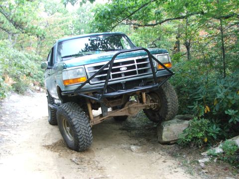 1996 Ford Bronco - Modified green Ford Bronco navigating rough terrain on a wooded trail.