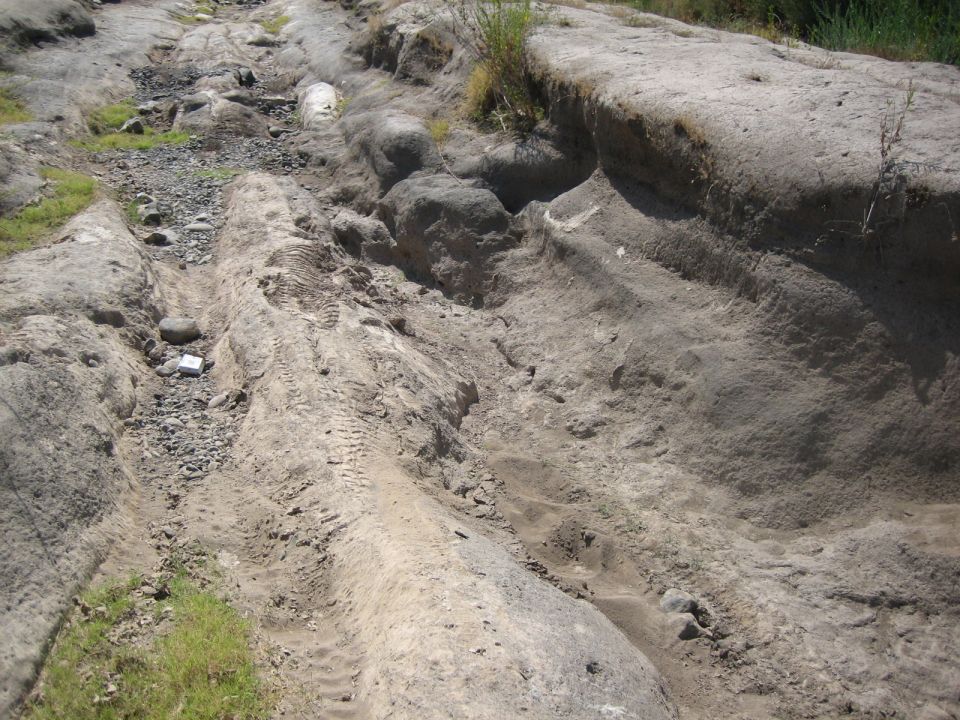 Tire tracks deeply imprinted in a dry, rocky wash with sparse vegetation and large boulders