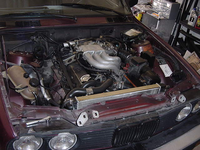 Maroon 1984 BMW 3 Series engine bay with V6 engine, radiator, and coolant reservoir visible