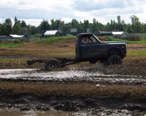 1979 Ford Bronco - A pickup truck chassis is driven through a muddy off-road course with trees and buildings in the background.