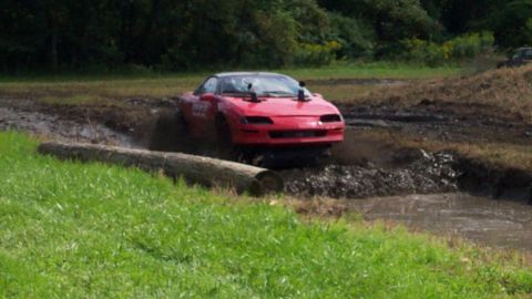 1979 Ford Bronco - A red sports car splashes through a muddy track, with a log obstacle in the foreground.