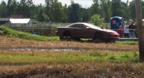 1979 Ford Bronco - A red sports car appears to be stuck in a muddy field with its wheels removed.