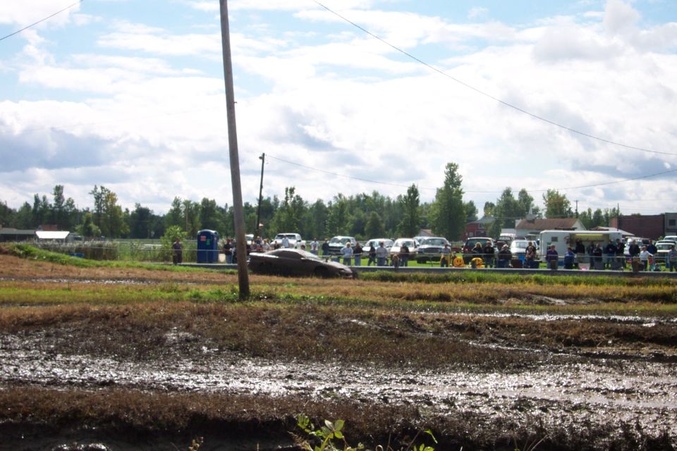 Muddy field with parked cars and spectators under a cloudy sky