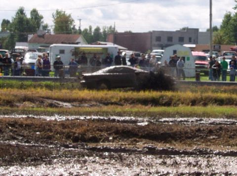 1979 Ford Bronco - A vehicle navigates a muddy track during an event, with onlookers observing from the sidelines.