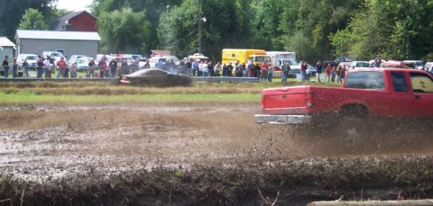 1979 Ford Bronco - A red pickup truck sprays mud during a race, with a brown car and onlookers in the background.