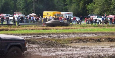 1979 Ford Bronco - A car is stuck in a mud pit during an event, with onlookers and emergency vehicles present.