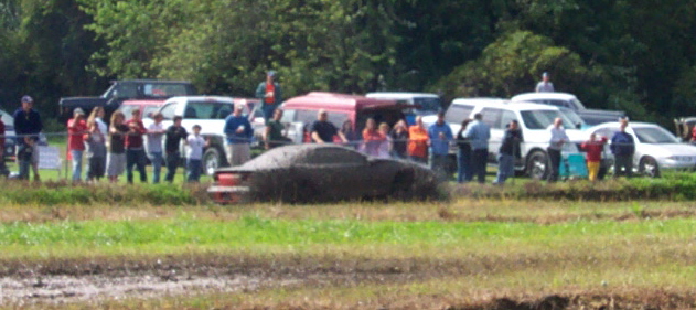 Car covered in mud, spectators watching from a grassy area with trees and other vehicles in the background