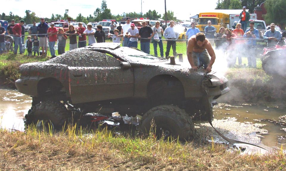 Mud-covered car stuck in a muddy field with a man on top, large tires, and a crowd watching