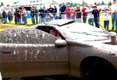 1979 Ford Bronco - Muddy car with occupants smiles at the camera while onlookers watch from a distance.