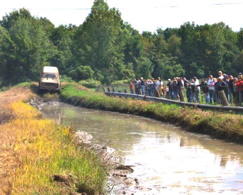 1979 Ford Bronco - A van navigates a muddy track as a crowd observes the event from a roadside barrier.