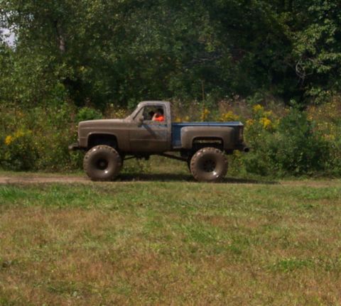 1979 Ford Bronco - A lifted pickup truck with oversized tires drives on a dirt path through a grassy field.