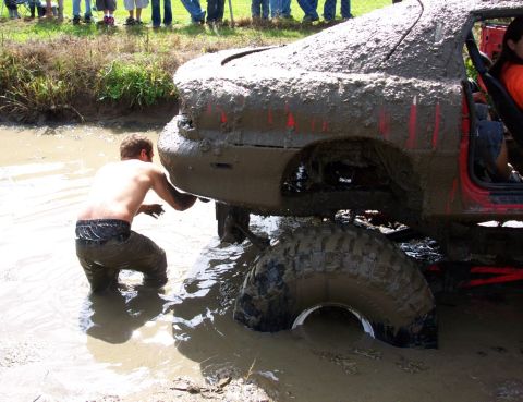 1979 Ford Bronco - A person wades in muddy water to inspect a heavily mud-caked red car stuck in a mire.