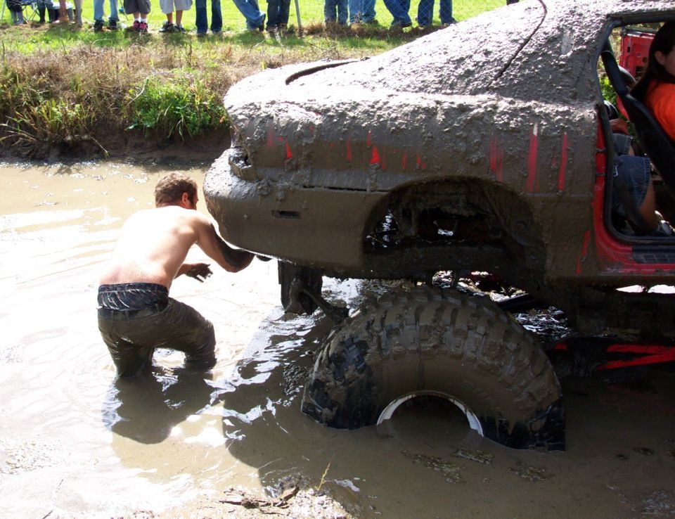Mud-covered red car with large tires stuck in water, a person is checking underneath