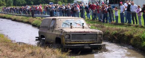 1979 Ford Bronco - A heavily mud-caked SUV plows through a muddy track, observed by a crowd of onlookers.