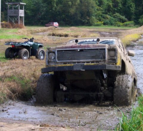 1979 Ford Bronco - A heavily mud-covered truck is stuck in a muddy track, with an ATV and a distant red SUV visible.