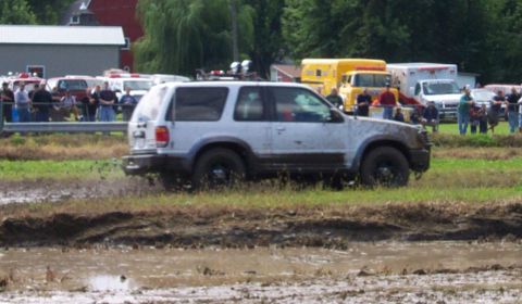 1979 Ford Bronco - A white SUV navigates a muddy track, while emergency vehicles and onlookers observe the scene.