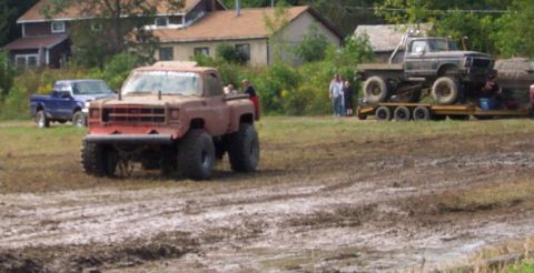 1979 Ford Bronco - A heavily modified red pickup truck with large tires is seen in a muddy field, with another truck on a trailer in the background.