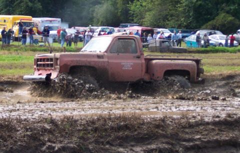1979 Ford Bronco - A red pickup truck is stuck deep in the mud during an event, with spectators and other vehicles in the background.