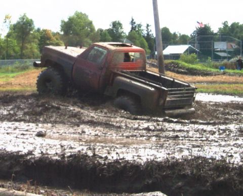1979 Ford Bronco - A lifted pickup truck is stuck deep in a muddy field, with trees and a baseball field in the background.